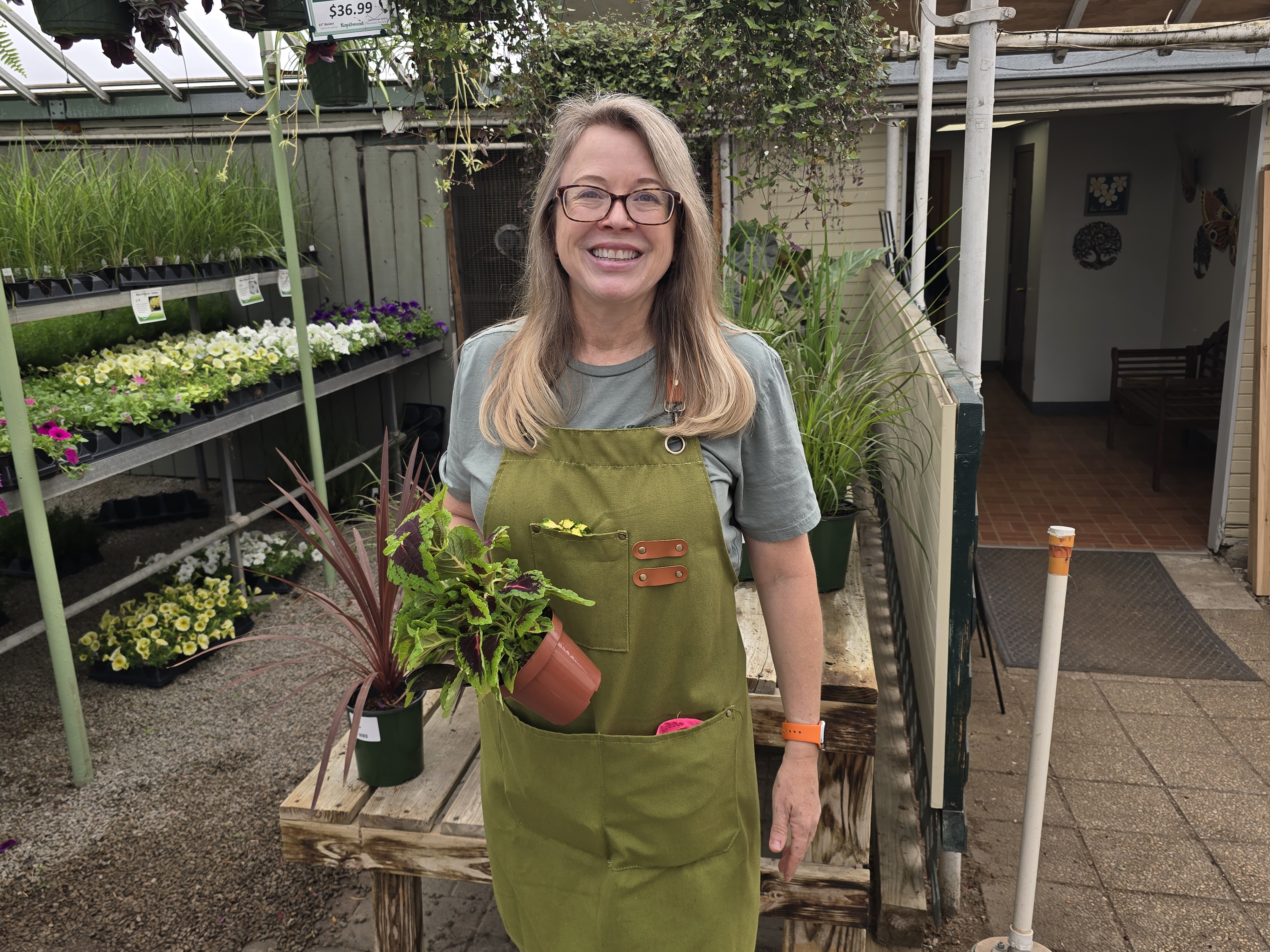 a woman in a green apron holding a potted plant in front of shelves of other plants that are for sale 